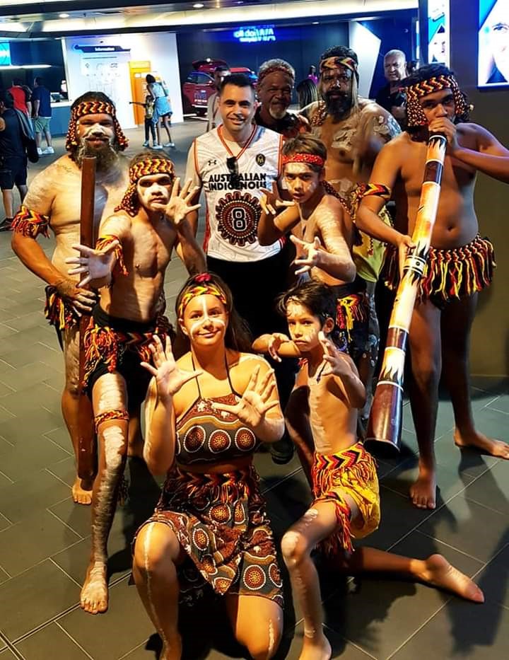 A group of eight boys and men and one woman of aboriginal heritage are grouped together in traditional dress and paint ready for a dance performance. These are the members of the Bindjareb Middars.